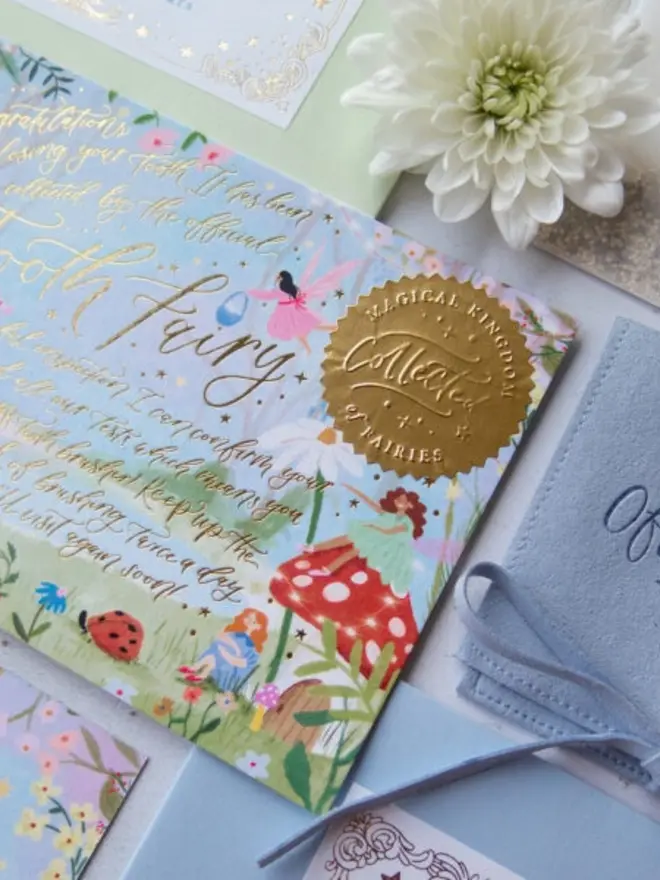 The gold "Collected" seal and fairy illustrations on the notecard. A pale blue suede pouch and a white chrysanthemum lie next to it.