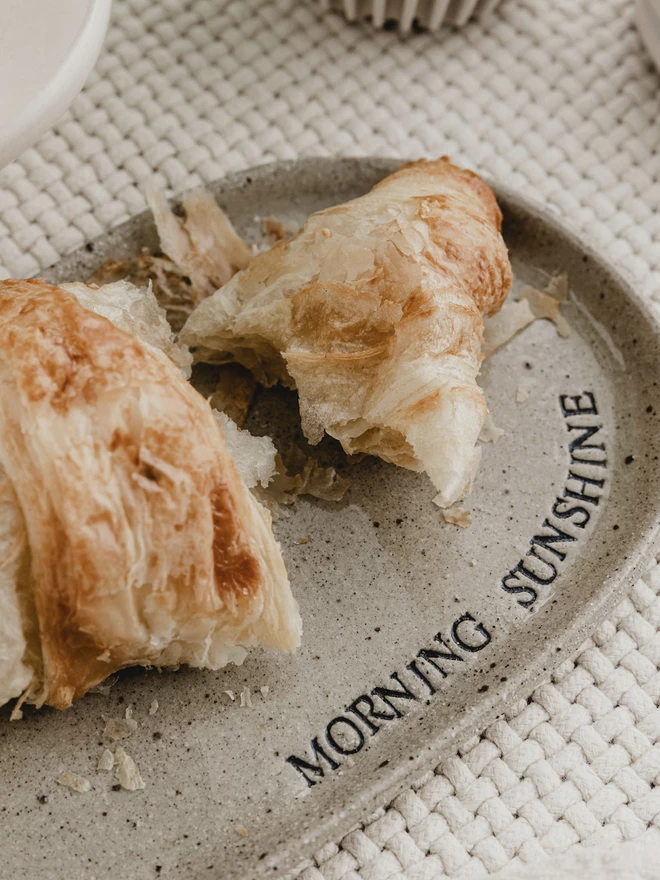 oval ceramic plate holding a croissant. Plate is stamped with the words 'morning sunshine'