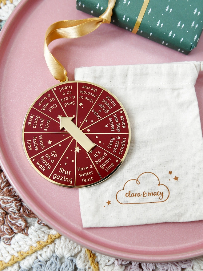 A deep red and gold Christmas decoration lays beside a cotton bag. It has 12 segments, each one with a different Christmas activity idea, and a golden arrow in the centre.