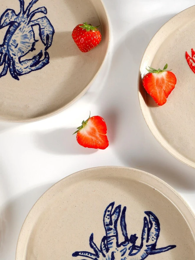 Handmade Ceramic Side Plate With Blue Lobster Design, a side plate with a blue lobster design sitting on a white table surface accompanied by a raspberry. 