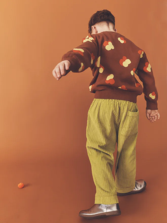 Back view of a child mid-kick against a burnt orange backdrop, showing the reverse of a Studio Koter chestnut jacquard jumper and chartreuse corduroy trousers with a rear patch pocket.