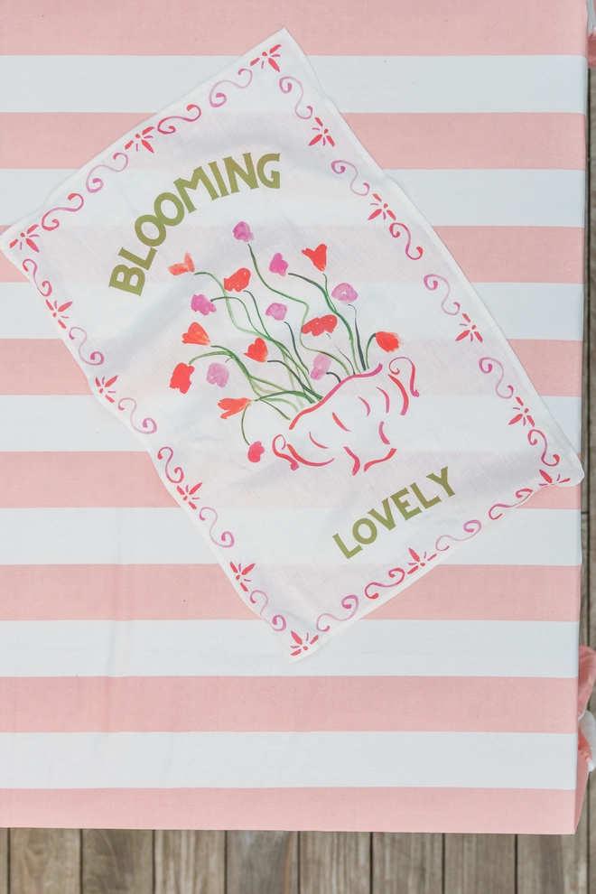 Linen teatowel with Blooming Lovely and floral pattern, laid against a pink and white striped tablecloth