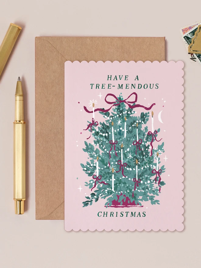 Pale pink Tree-mendous Christmas card with a scalloped edge and a candlelit Christmas tree tied with bows, lettered 'Have a tree-mendous Christmas', on a kraft envelope beside a gold pen