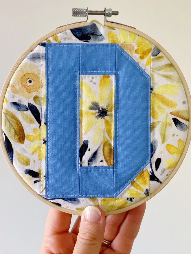 Personalised quilted hoop on a floral yellow background with a blue letter 'D.'