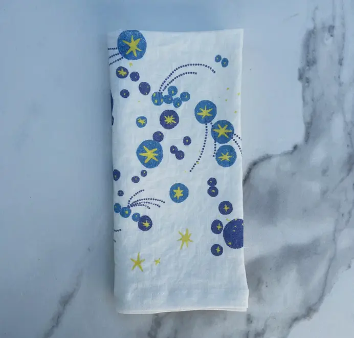 Solaris Napkins In White Or Blue. Off-white cloth napkin with blue and yellow starburst and orbital patterns. 