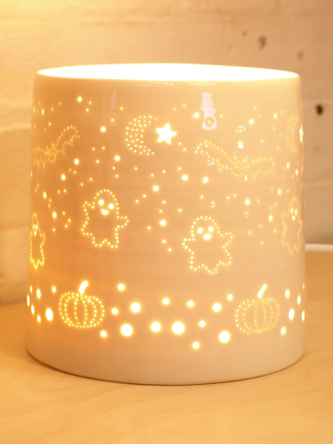 A ceramic cylindrical lamp, made from porcelain, featuring a design pierced into the clay of ghosts, pumpkins bats and stars.