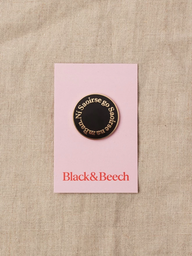 A black round enamel pin with the words Ní Saoirse go Saoirse na mBan written around the edge in gold lettering on a pink backing card 
