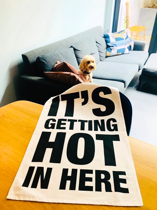 London Drying It's Getting Hot in Here black screen printed text on white tea towel laying on table with dog on sofa in background