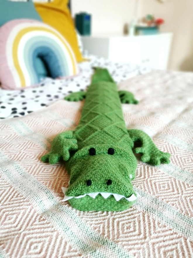 A close up of Claude the Crocodile long hot water bottle pictured laying diagonally across a double bed. Colourful rainbow cushions can be seen in the background.