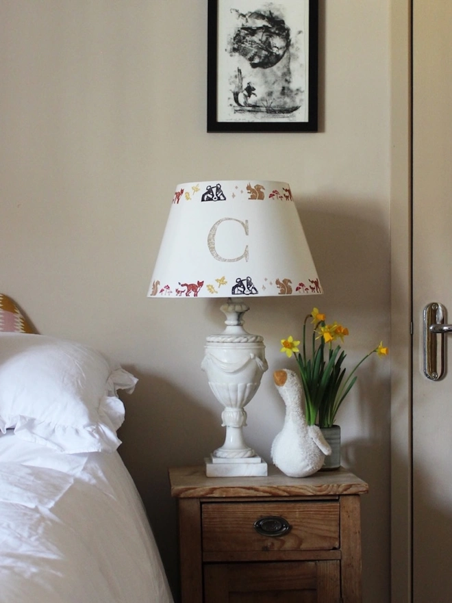 A cosy children's bedroom with a rustic wooden nightstand featuring a personalised lampshade with the letter "C" and a countryside pattern. A plush goose, daffodils, and white bedding add charm. Neutral empire light shade with pretty animal details, ideal for kids bedroom design and personalised kids gift.