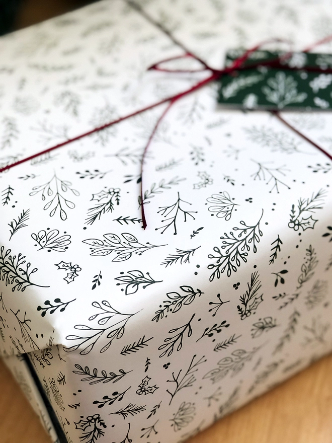 A gift wrapped in white wrapping paper with a Christmas botanical design lays on a wooden floor.