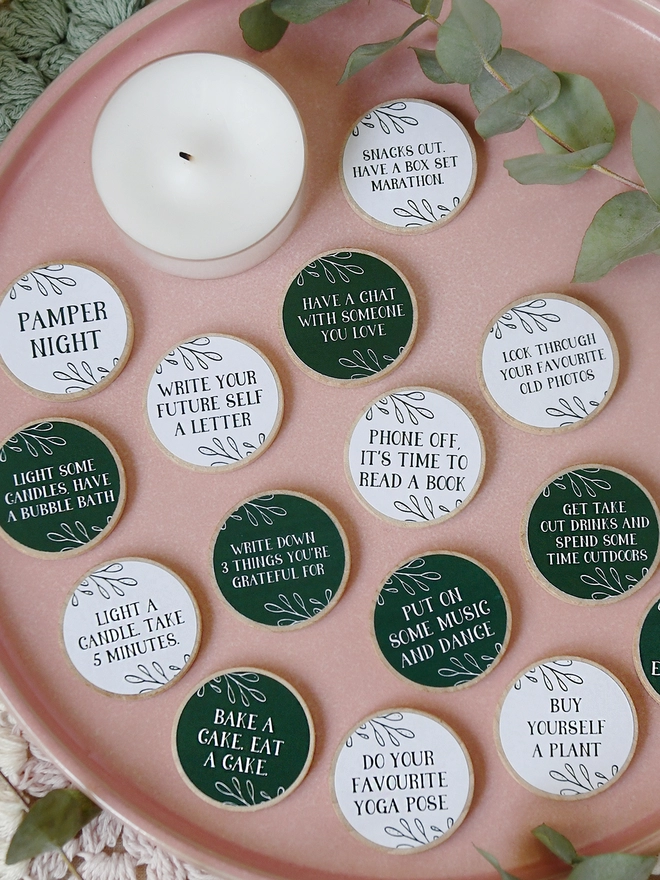 Fifteen wooden tokens with dark green and white labels, each with a self care idea printed on, lay on a pink plate.