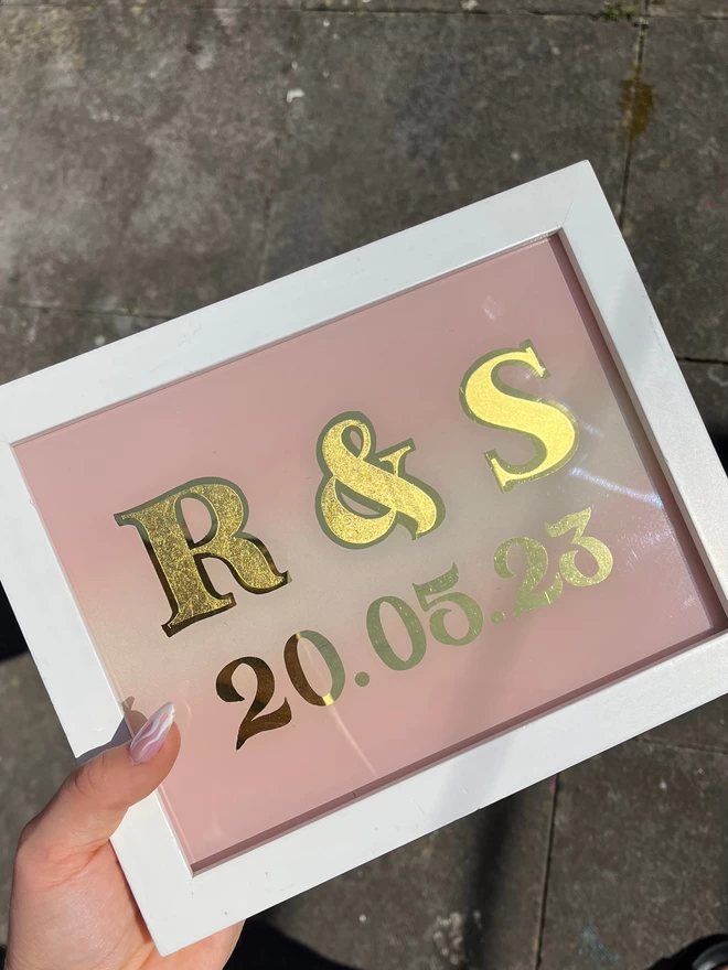 White framed gilded sign with a pink background and text spelling 'R&S 20.05.23'