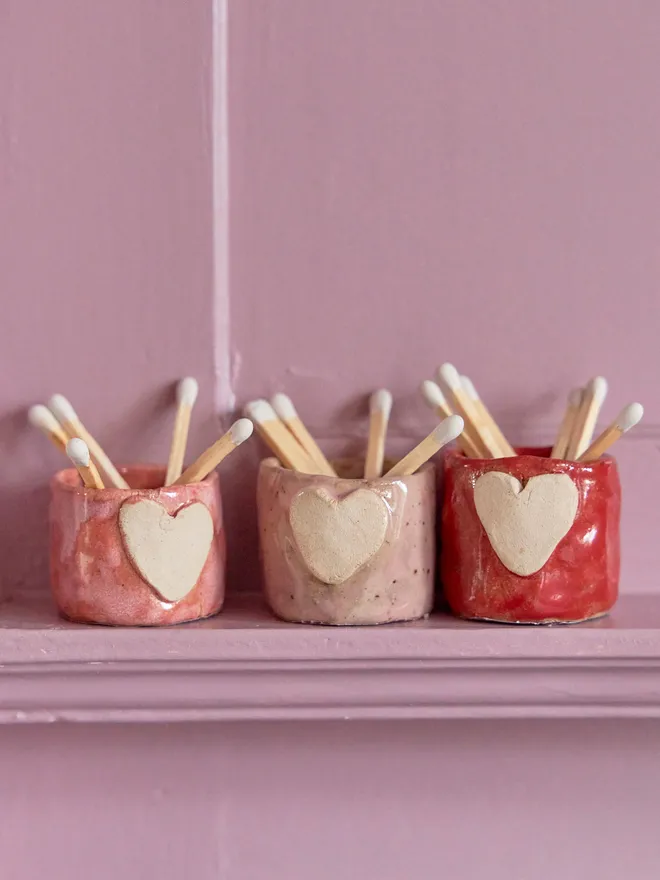Mini ceramic match holder in red and pink on a pink shelf with white matches inside.