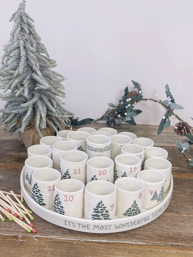 alternative advent tray with 24 numbered candle holders on a rustic table top with christmas tree and matches