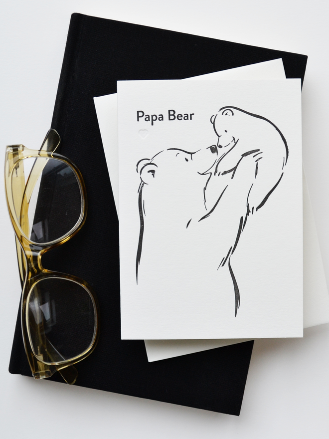 Black and white greeting card resting on a black book with reading glasses.  Card features bear characters, simple illustration of 'papa bear' holding aloft baby bear with words that read Papa Bear