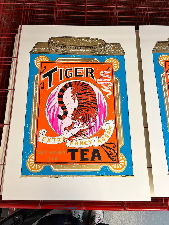 Tiger Tea Tin Fine And Fancy, a colourful tea design on a white screenprint. It sits on a studio rack next to more screenprints. 