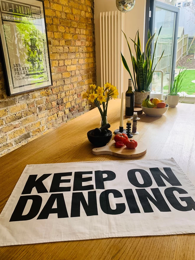 London Drying Keep on Dancing black screen printed text on white tea towel laying flat on timber kitchen island with plant, daffodils in vase, fruit bowl, chopping board with vegetables in background