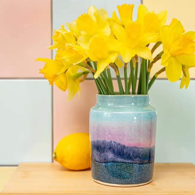 Landscape Medium Vase, a colourful vase sitting on a wooden surface against a patterned backdrop. It is accompanied by some flowers. 