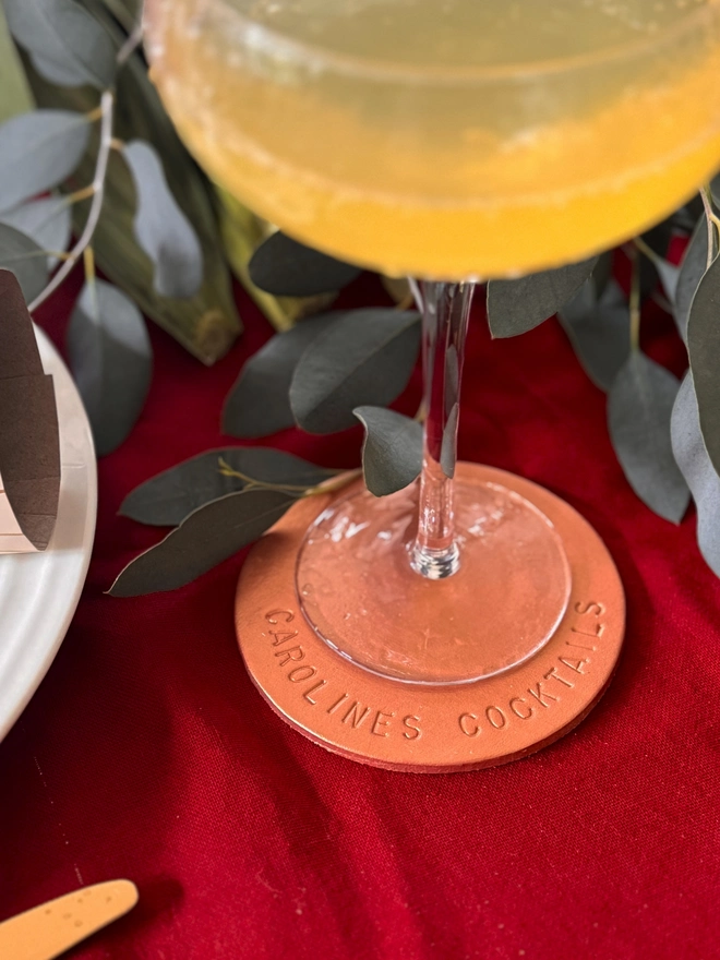 rose gold leather coaster, with caroline's cocktails stamped around the edge. A bucks fizz resting on the coaster.