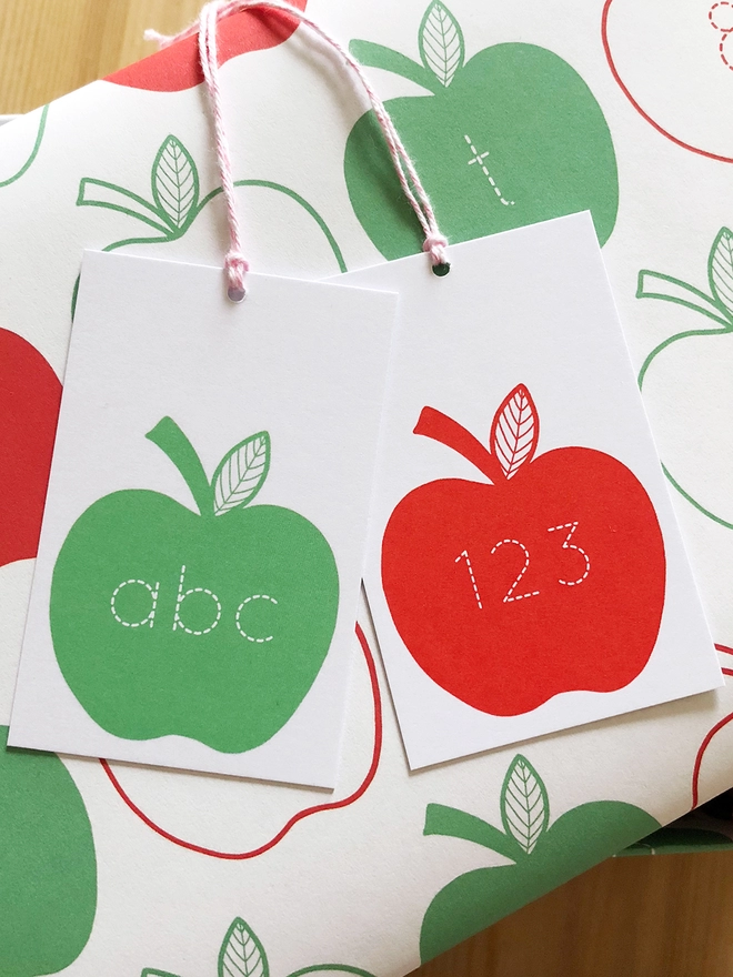 A gift wrapped in red and green apple wrapping paper, with letters and numbers inside each apple, is on a wooden surface.