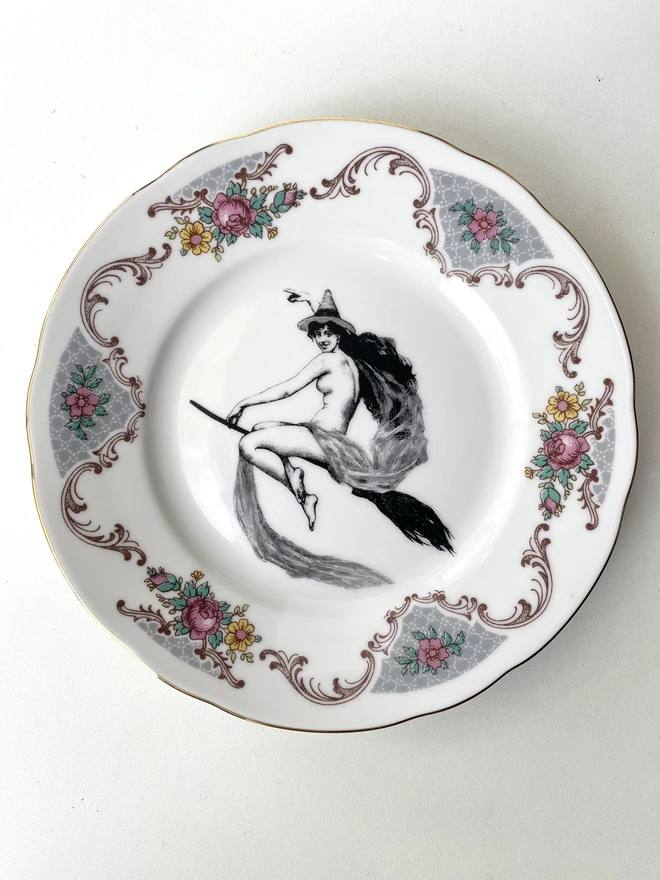 vintage plate with an ornate border, with a printed vintage illustration of a witch on a broomstick in the middle 