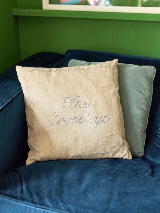 Embroidered linen cushion with dusty blue stitching for a family name and date, on a blue sofa with a light blue cushion behind, against a bright green wall