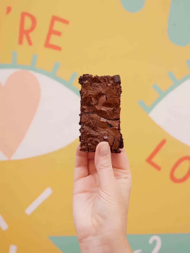 Gluten free classic fudge brownie slice being held by a hand against a colourful background