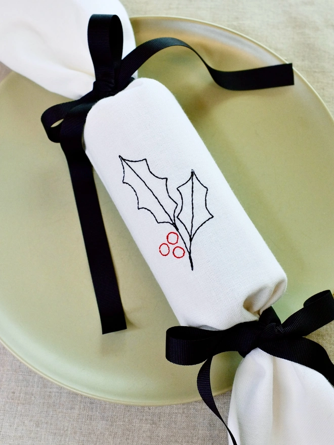 White cotton Christmas cracker with embroidered holly design and black ribbon bows 