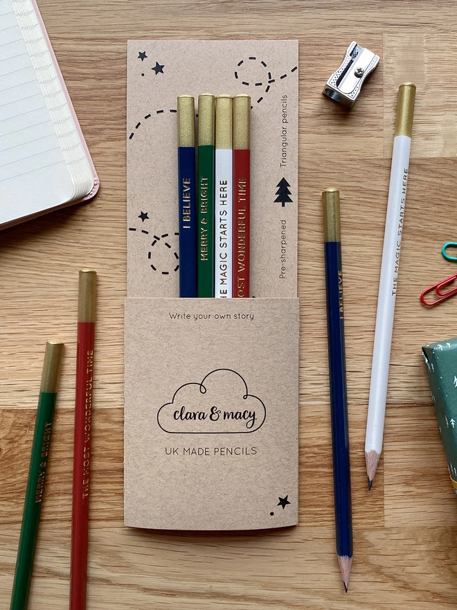 Four festive coloured pencils with gold writing along the side are tucked into cardboard packaging on a wooden desk. Loose pencils and stationery items lay beside them.