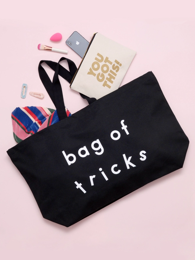 model holding out an oversized black canvas tote bag with the words bag of tricks laying on a pink backdrop with items spilling out