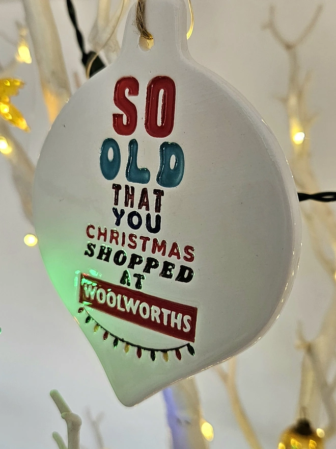 A handmade flat white ceramic bauble which measures 10cm by 8cm with So old that you Christmas shopped at Woolworths written on it in colourful glazes and retro fonts with a little string of colourful fairy lights stamped into the clay.