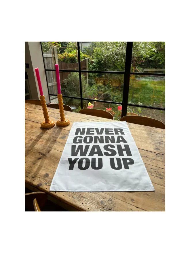 Never Gonna Wash You Up screen printed tea towel laying on wooden table next to candlesticks with window/garden view in background