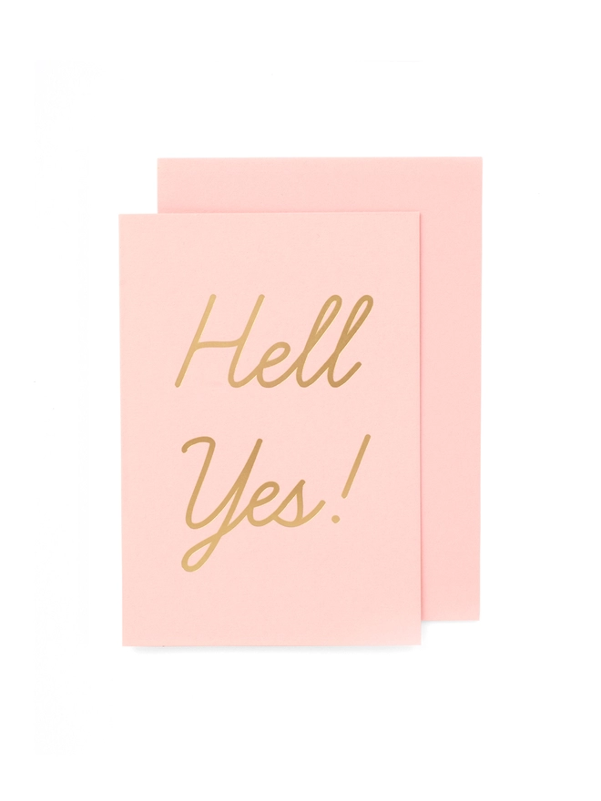 A pink greeting card that says 'Hell Yes!' in gold foil
