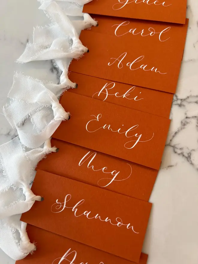 Personalised Calligraphy Rust Orange Place Card, orange place cards with white chiffon ribbons grouped together on a white surface. 