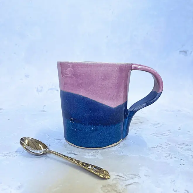 Landscape Standard Mug, a colourful mug placed on a stone surface. It is accompanied by a spoon. 