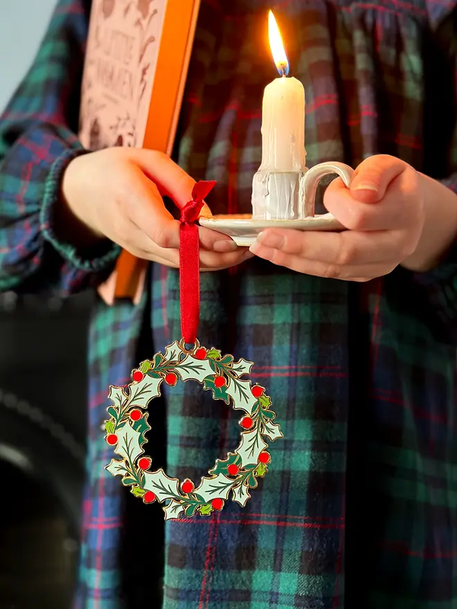 A child wearing a tartan dress holds a vintage-style enamel Christmas wreath ornament with green holly leaves and red berries, hanging from a red ribbon, alongside a small white candleholder with a lit candle.