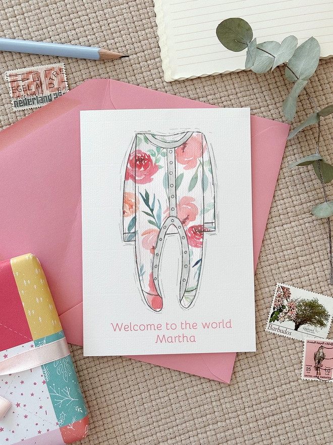 An illustrated new baby greetings card with a pink onesie design lays on a pink envelope beside various stationery items.