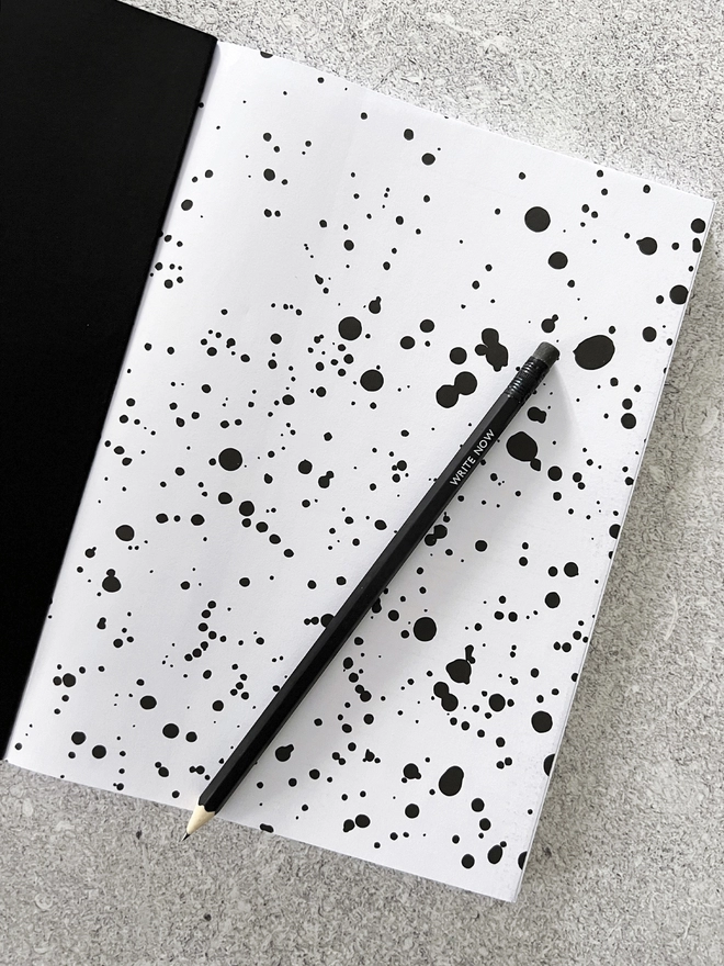 Notebook open to the first page with the black and white inkwell pattern with a black pencil laid on top. 