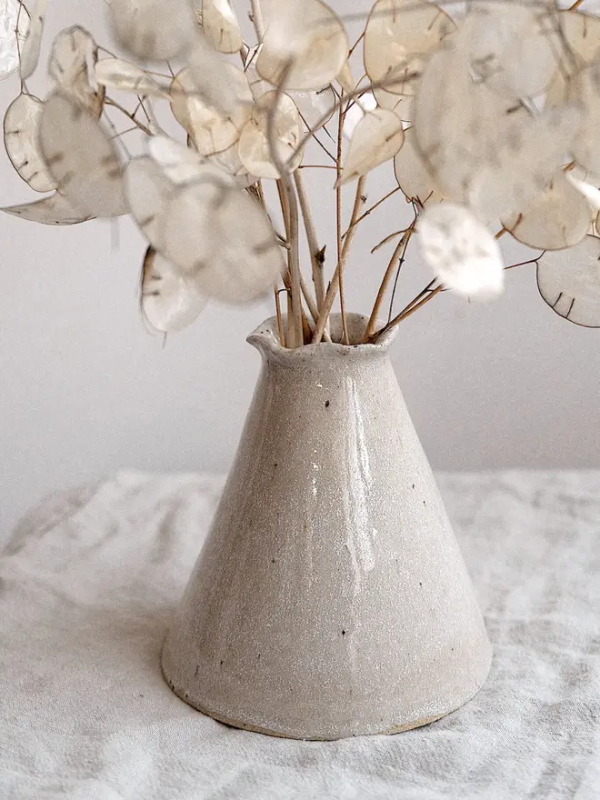 Maia Bud Vase In Sea Salt Glaze, with plant in it, placed on a table cloth against a white background. 