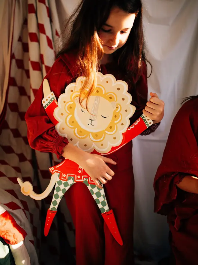 Extra Large Lion Boy Wall Puppet. a young girl in a red velvet outfit holds the extra large lion boy wall puppet close to her chest, smiling as sunlight catches her hair. the lion puppet has a cream and gold mane, red jacket with white trim, and green and white checked leggings ending in red boots, with jointed wooden limbs. the warm, golden lighting and soft focus on a second child nearby give the scene a nostalgic, storybook feel.