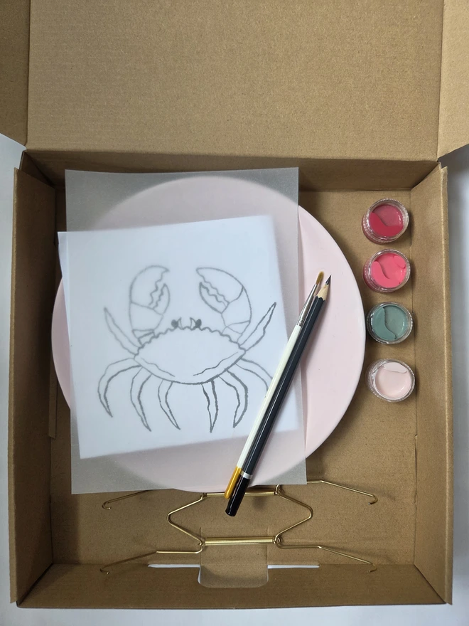 cardboard box containing the contents of the crab plate painting kit - plate, design template, brush, pencil, paints and wall hook. 
