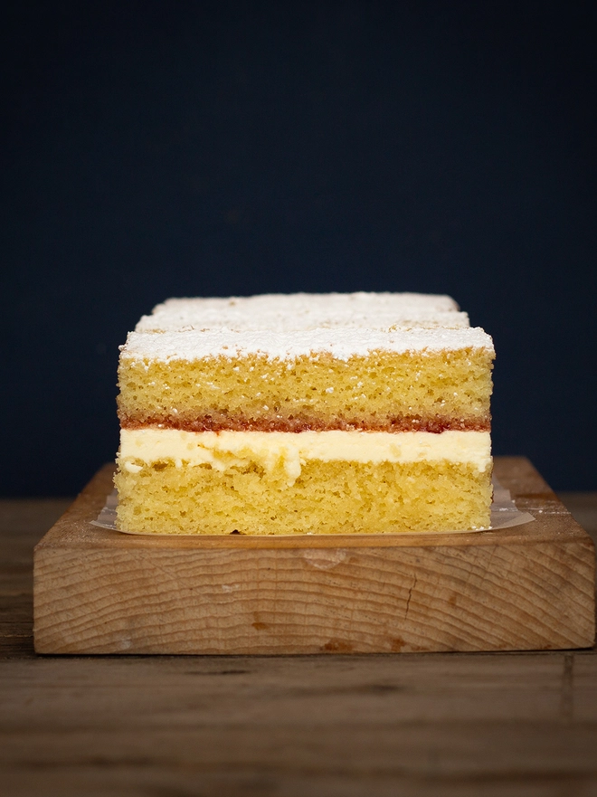 Side profile of the Victoria Sponge Cake showing a bright strawberry red jam on top of a creamy butter cream, sandwiched between 2 soft sponge cakes with a showering of icing sugar on top.