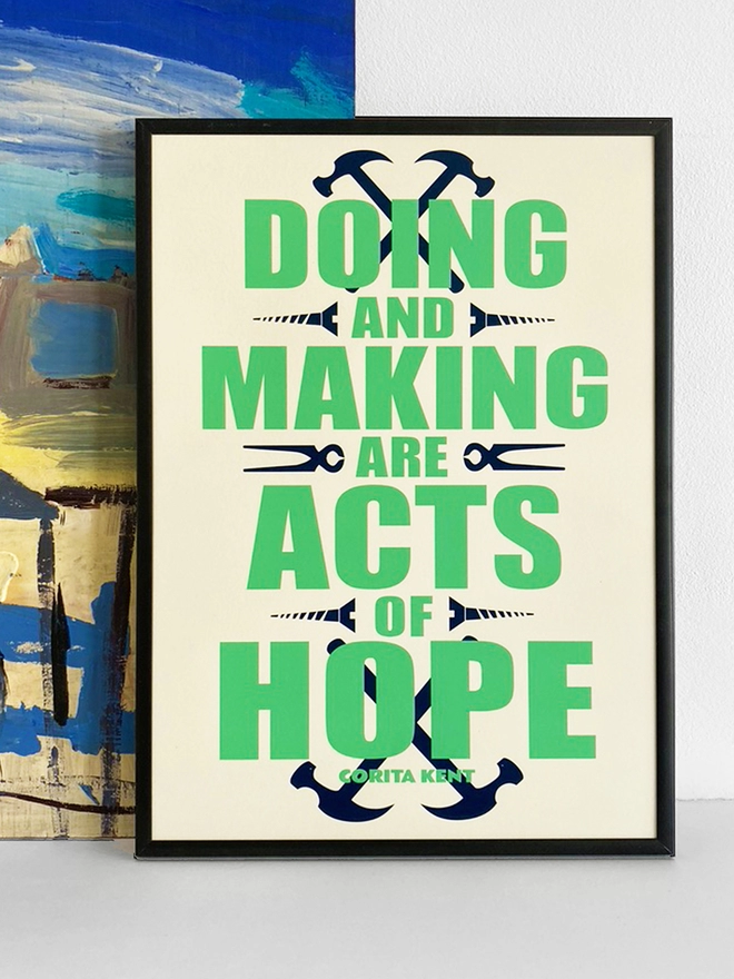 Framed green and blue typographic print. The Corita Kent quote reads "Doing and making are acts f hope." Navy blue tools are placed around the green text. The print rests against a blue and yellow abstract painting.