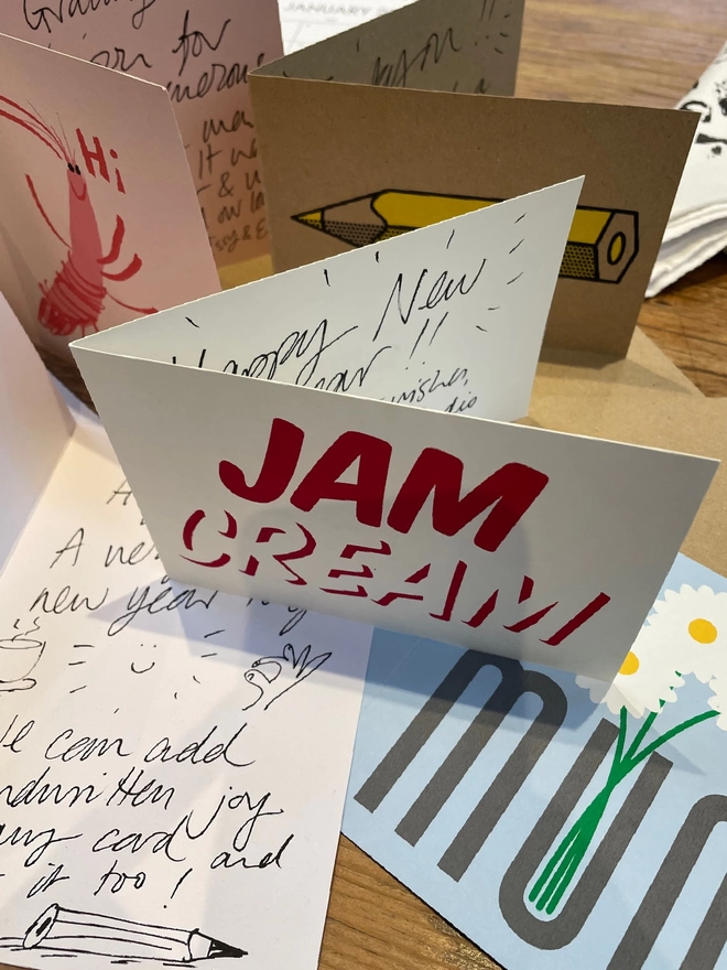An array of colourful bold greetings card are stood and laid out with glimpses of energetic handwritten messages, Jam Cream typography, the word Mum with flowers poking out, A prawn says hi!, a graphic yellow pencil. Exuding joy and life!