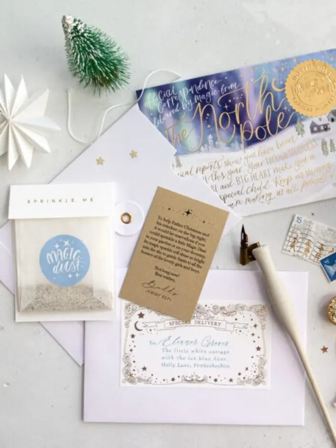 A Magic Dust bag, a kraft instruction card and a white envelope with a personalised "Special Delivery" label addressed to Eleanor Groves, in front of the northern lights North Pole notecard. A white dip pen, vintage stamps and a miniature frosted tree are arranged beside them.