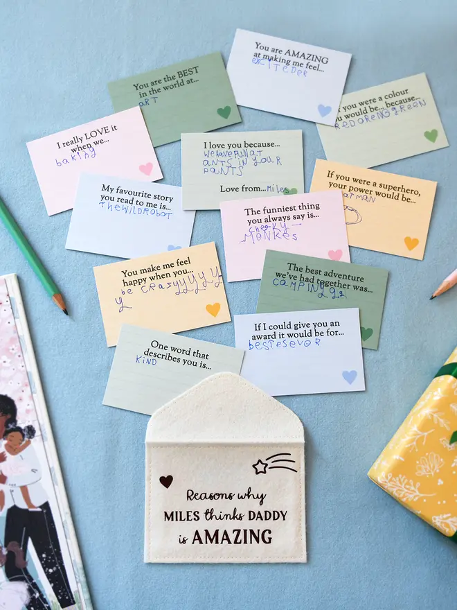 A flat lay of a handmade cream felt envelope with the words "Reasons why MARTHA thinks DADDY is AMAZING" on the front, surrounded by 12 colourful prompt cards filled in by a child with handwritten messages and drawings. The cards include prompts like "You are the BEST in the world at..." and "If I could give you an award...". Also visible are coloured pencils, a storybook, on a blue surface.