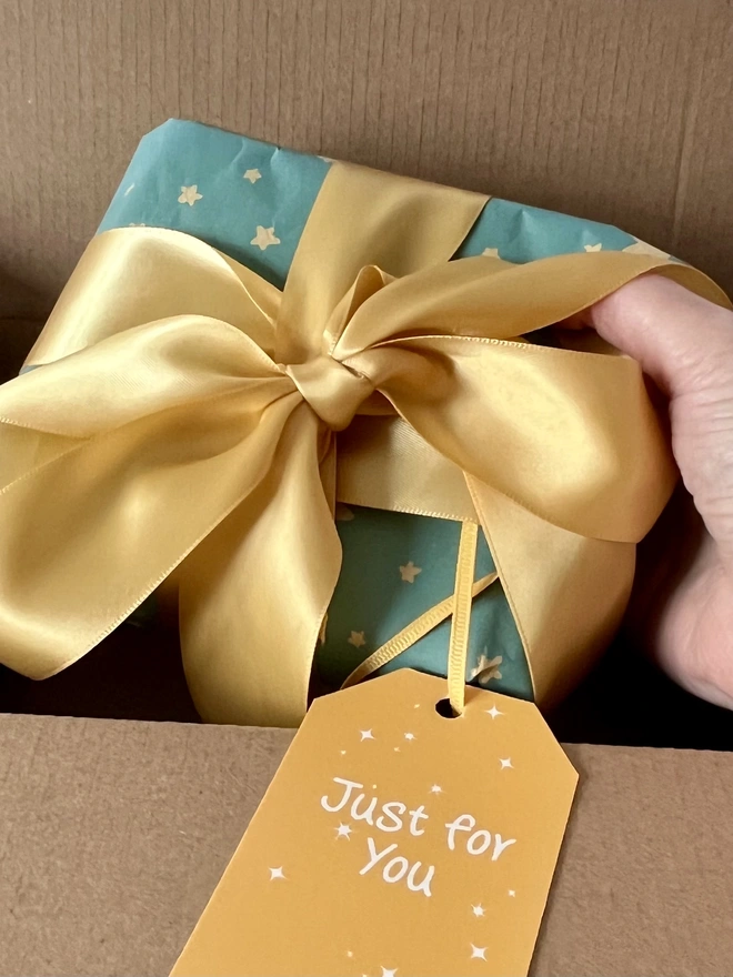 Hand placing small square parcel into a brown box for posting. The parcel is gift wrapped in blue paper with gold stars & large gold bow. A yellow gift tag says 'Just for You' & has small white stars scattered over it. 