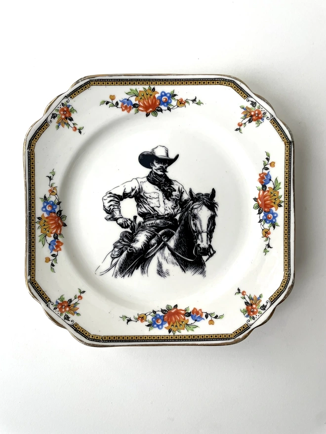 vintage plate hanging on a white wall, it has a decorative border, with a printed vintage illustration of a cowboy in the middle 