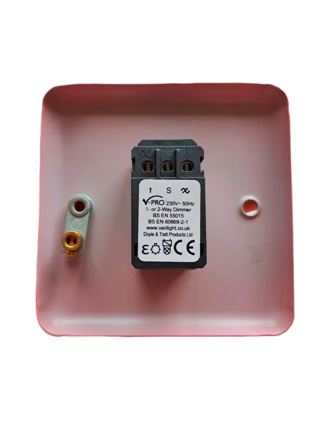 The back of a pink steel metal epoxy coated light switch plate, there is a black rectangle LED dimmer switch module in the centre of the switch plate, the module has three round holes at it’s base for wires and a white label with V Pro, 230v – 50Hz 1 or 2 way Dimmer, BS EN 55015, BS EN 60669 – 2- 1 www.varilight.co.uk, Doyle & Tratt products Ltd written one it, there are two screw holes in the light switch, the hole on the left also has an earthing screw on it.
